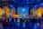 A large indoor space with people seated on the floor, facing a central stage where a performer is surrounded by colorful lighting. The room is decorated with large plants and nature-themed murals. Visuals on a big screen enhance the atmosphere.