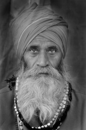 A black and white portrait featuring an older man with a thick beard and a turban. He wears a necklace with distinct beads and his expression is contemplative. The background is softly blurred, focusing attention on the man&rsquo;s striking features.