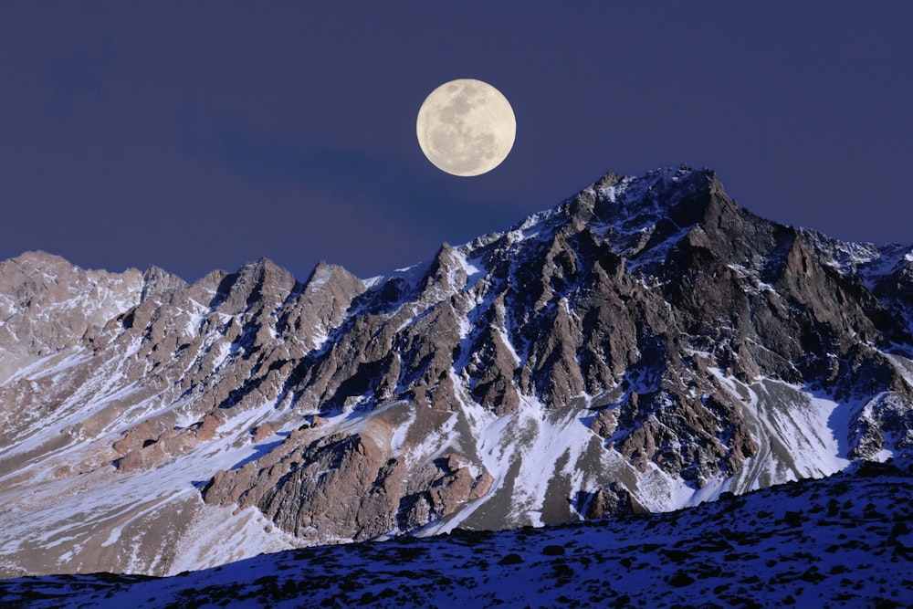 a full moon rises over a snowy mountain range