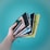 A hand holding several colorful notebooks with elastic closures against a turquoise backdrop. The notebooks are in various colors including black, blue, green, and yellow.
