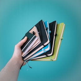 A hand holding several colorful notebooks with elastic closures against a turquoise backdrop. The notebooks are in various colors including black, blue, green, and yellow.