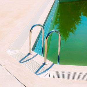 A swimming pool with a distinctly green-tinted water is bordered by a light-colored deck. The pool features a chrome pool ladder with two handles that cast shadows on the surrounding surface. The tile lining the pool's edge is a blue color, contrasting with the green water.