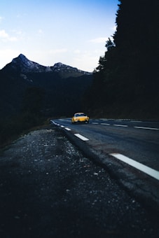 A winding road cuts through a mountainous landscape with lush trees. A bright yellow car drives along the road, creating a vivid contrast against the darker, overcast scenery. In the background, rugged mountains rise into the sky, partially shrouded in shadow. The overall scene conveys a sense of solitude and adventure as the car journeys along the empty road.