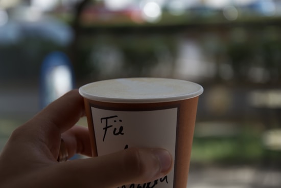 A hand holds a brown paper coffee cup with a white lid, featuring handwritten text in black ink. The background is blurred, suggesting an outdoor setting with greenery and out-of-focus objects.