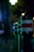A series of bollards lined up along a street at night, each with a reflective band. The scene is dimly lit with bright, blurred lights in the background, creating a moody urban setting. Raindrops are visible on the surface of the bollards.