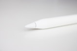 A close-up view of the tip of a stylus pen set against a light, neutral background. The pen is sleek and predominantly white, with a minimalistic design that emphasizes its smooth surface and tapered point.