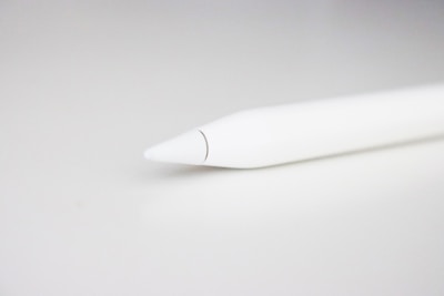 A close-up view of the tip of a stylus pen set against a light, neutral background. The pen is sleek and predominantly white, with a minimalistic design that emphasizes its smooth surface and tapered point.