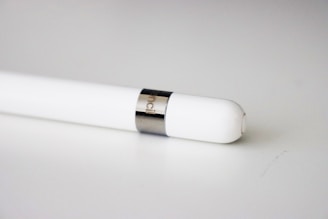 A white stylus pen with a smooth, sleek design is placed on a light-colored surface. It features a metallic band near the tip, and 'pencil' is engraved on the band.