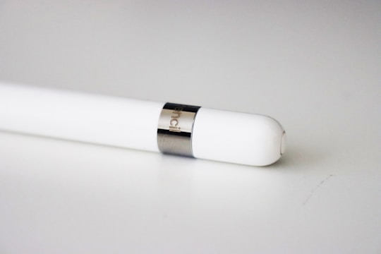 A white stylus pen with a smooth, sleek design is placed on a light-colored surface. It features a metallic band near the tip, and 'pencil' is engraved on the band.