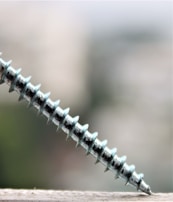 A metallic screw is positioned diagonally, partially resting on a surface with a blurred background. The screw's threads are prominently visible and reflect a slight sheen.