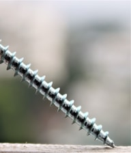 A metallic screw is positioned diagonally, partially resting on a surface with a blurred background. The screw's threads are prominently visible and reflect a slight sheen.