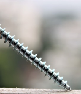 A metallic screw is positioned diagonally, partially resting on a surface with a blurred background. The screw's threads are prominently visible and reflect a slight sheen.