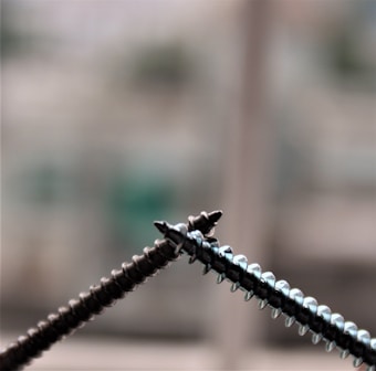 Close-up view of two intersecting metal screws set against a blurred background. The screws are metallic, showing intricate threads and sharp points.