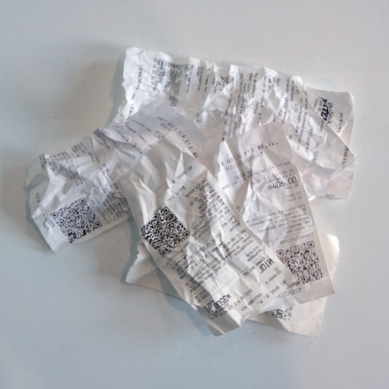 Crumbled and wrinkled paper receipts are strewn across a smooth white surface. The receipts contain printed text and QR codes, indicating they are from various transactions.