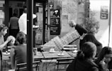 A bustling café scene captures several people seated outdoors at wooden tables, enjoying drinks and conversation. In the background, a man is carrying a large stack of water bottles toward a bar entrance. The café has a rustic charm with a chalkboard menu displayed on the wall.