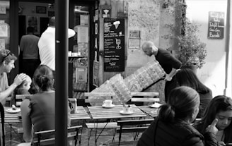 A bustling café scene captures several people seated outdoors at wooden tables, enjoying drinks and conversation. In the background, a man is carrying a large stack of water bottles toward a bar entrance. The café has a rustic charm with a chalkboard menu displayed on the wall.