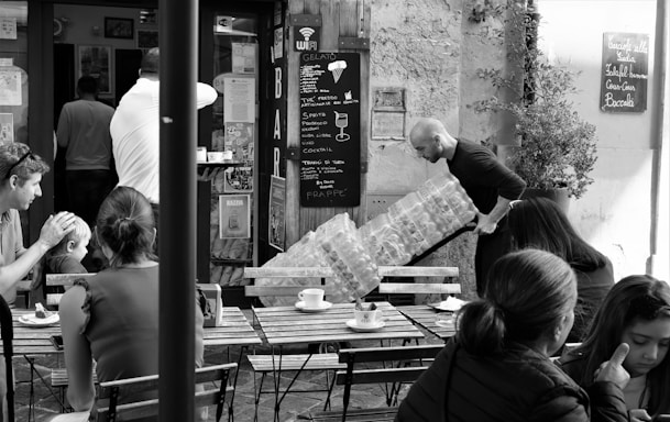 A bustling café scene captures several people seated outdoors at wooden tables, enjoying drinks and conversation. In the background, a man is carrying a large stack of water bottles toward a bar entrance. The café has a rustic charm with a chalkboard menu displayed on the wall.