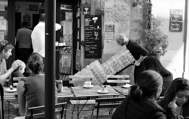 A bustling café scene captures several people seated outdoors at wooden tables, enjoying drinks and conversation. In the background, a man is carrying a large stack of water bottles toward a bar entrance. The café has a rustic charm with a chalkboard menu displayed on the wall.