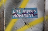 A textured wall with cracks features a prominent sticker displaying the words 'Life Requires Movement' in bold, white letters. The background is a mix of gray and yellow spray paint, creating a dynamic, urban feel.