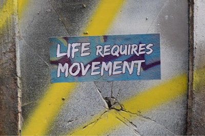A textured wall with cracks features a prominent sticker displaying the words 'Life Requires Movement' in bold, white letters. The background is a mix of gray and yellow spray paint, creating a dynamic, urban feel.