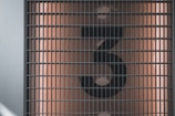 A large, black, stylized number 3 is displayed behind a metal grid. The background is a muted, warm beige color with light reflections visible.