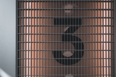 A large, black, stylized number 3 is displayed behind a metal grid. The background is a muted, warm beige color with light reflections visible.