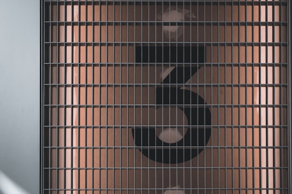 A large, black, stylized number 3 is displayed behind a metal grid. The background is a muted, warm beige color with light reflections visible.