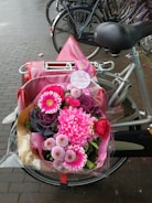 A colorful bouquet of pink flowers, including gerberas and roses, is placed on the back of a bicycle with a pink bag. The flowers are wrapped in clear cellophane and the bike is parked on a wet brick pavement.