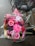 A colorful bouquet of pink flowers, including gerberas and roses, is placed on the back of a bicycle with a pink bag. The flowers are wrapped in clear cellophane and the bike is parked on a wet brick pavement.