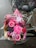 A colorful bouquet of pink flowers, including gerberas and roses, is placed on the back of a bicycle with a pink bag. The flowers are wrapped in clear cellophane and the bike is parked on a wet brick pavement.