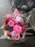 A colorful bouquet of pink flowers, including gerberas and roses, is placed on the back of a bicycle with a pink bag. The flowers are wrapped in clear cellophane and the bike is parked on a wet brick pavement.