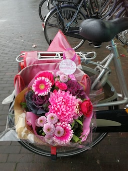 A colorful bouquet of pink flowers, including gerberas and roses, is placed on the back of a bicycle with a pink bag. The flowers are wrapped in clear cellophane and the bike is parked on a wet brick pavement.