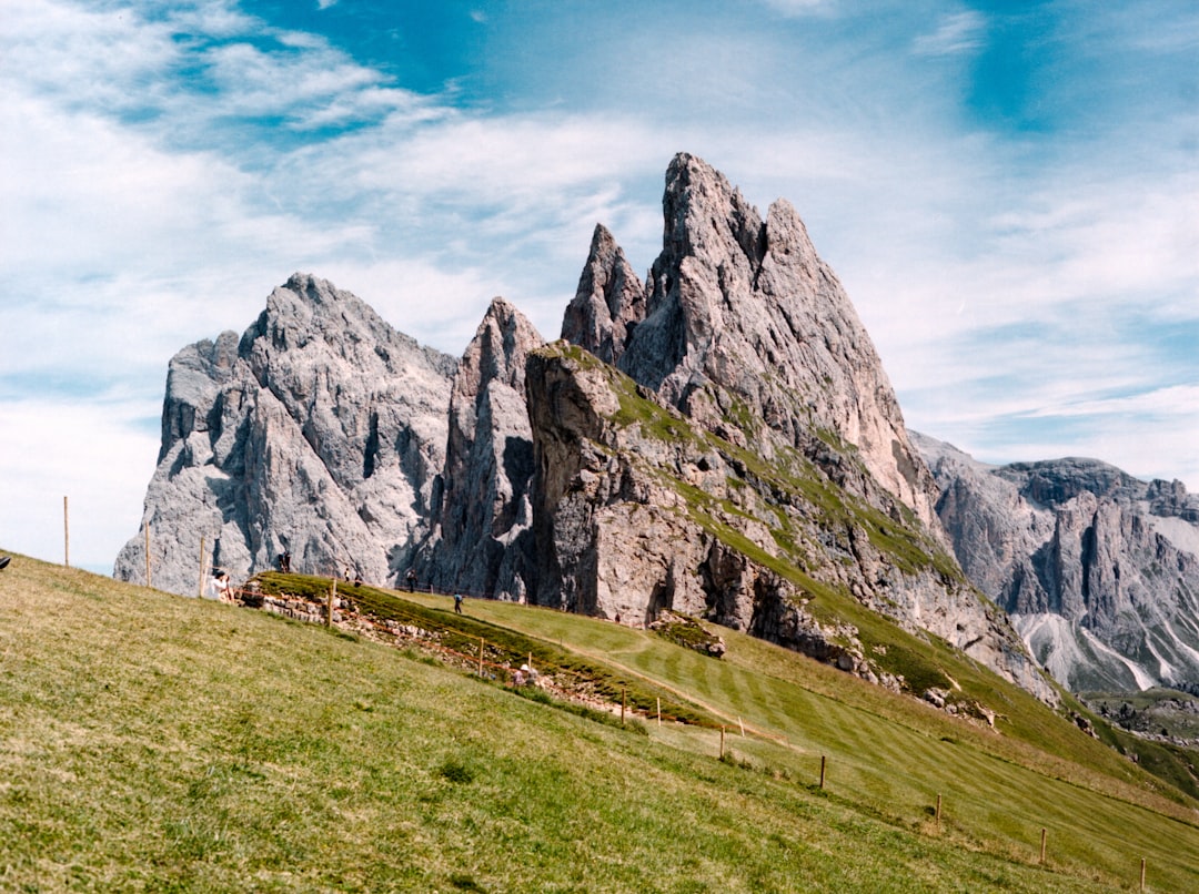 green grass field near gray rocky mountain under blue and white sunny cloudy sky during daytime