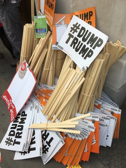 A pile of protest signs and placards leaning against a wall. The signs, primarily in black and white with bold lettering, prominently display the hashtag '#DUMP TRUMP' and some variations. Some signs are orange with similar messaging. Wooden sticks for holding the signs are bundled together alongside the placards.