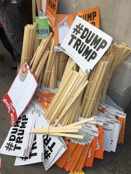 A pile of protest signs and placards leaning against a wall. The signs, primarily in black and white with bold lettering, prominently display the hashtag '#DUMP TRUMP' and some variations. Some signs are orange with similar messaging. Wooden sticks for holding the signs are bundled together alongside the placards.