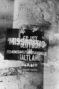 A black and white photo of a weathered poster on a textured wall. The poster contains several German words in bold, overlapping text, some of which are obscured. The background shows a gritty texture with signs of wear and tear.