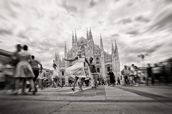 A couple dances joyfully in a bustling square with a prominent Gothic cathedral in the background. The scene is lively, with people walking around, adding to the vibrant atmosphere. The sky is overcast, enhancing the contrast of the architecture and the movement of the dancers.