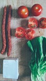 A wooden cutting board holds an arrangement of fresh ingredients including whole and halved tomatoes, two long sausages, a block of white cheese, and a bunch of green leafy lettuce.