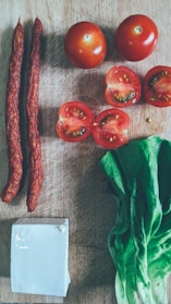 A wooden cutting board holds an arrangement of fresh ingredients including whole and halved tomatoes, two long sausages, a block of white cheese, and a bunch of green leafy lettuce.