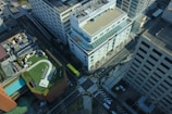 An aerial view of a cityscape featuring multiple high-rise buildings, with prominent white and gray structures. One building has a green rooftop garden with seating arrangements and pathways. There are visible roads with crosswalks and a few vehicles, including a yellow bus.