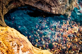 A small pool of clear water surrounded by textured stone. Pebbles in various colors are visible through the transparent water, creating a vibrant and natural mosaic. The surface of the water reflects light softly, creating gentle ripples.