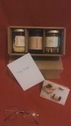 A cardboard box containing three jars labeled with different flavors is positioned against a red background. One jar is filled with crunchy ingredients, another with almond dark chocolate, and the last with biscuit sticks. A greeting card with 'Happy Birthday' written on it is placed in front of the box. Additionally, a pair of round eyeglasses and a small card featuring an image of a dessert next to a jar are arranged beside the greeting card.