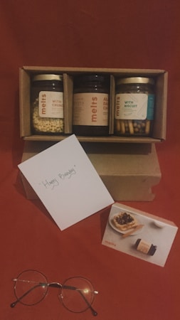 A cardboard box containing three jars labeled with different flavors is positioned against a red background. One jar is filled with crunchy ingredients, another with almond dark chocolate, and the last with biscuit sticks. A greeting card with 'Happy Birthday' written on it is placed in front of the box. Additionally, a pair of round eyeglasses and a small card featuring an image of a dessert next to a jar are arranged beside the greeting card.