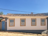 A single-story house with white walls and four windows outlined in yellow and blue. It has a traditional, textured tile roof. A light blue door is situated on the left side next to a black lantern fixture. The structure is set on a cobblestone street with clear blue skies in the background, and a few trees are visible behind the property.