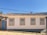 A single-story house with white walls and four windows outlined in yellow and blue. It has a traditional, textured tile roof. A light blue door is situated on the left side next to a black lantern fixture. The structure is set on a cobblestone street with clear blue skies in the background, and a few trees are visible behind the property.