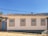 A single-story house with white walls and four windows outlined in yellow and blue. It has a traditional, textured tile roof. A light blue door is situated on the left side next to a black lantern fixture. The structure is set on a cobblestone street with clear blue skies in the background, and a few trees are visible behind the property.