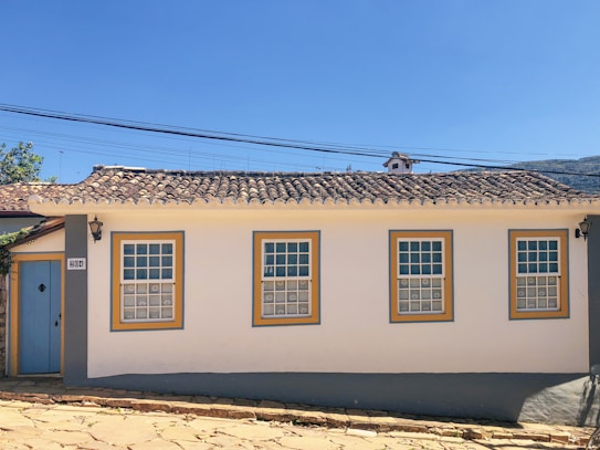 A single-story house with white walls and four windows outlined in yellow and blue. It has a traditional, textured tile roof. A light blue door is situated on the left side next to a black lantern fixture. The structure is set on a cobblestone street with clear blue skies in the background, and a few trees are visible behind the property.