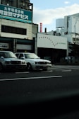 Several taxis are parked alongside a street in front of a commercial building with signage in Japanese. The sky is partially cloudy, and the overall setting gives an urban feel.