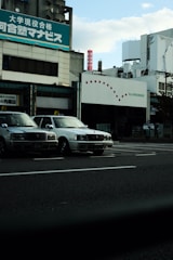 Several taxis are parked alongside a street in front of a commercial building with signage in Japanese. The sky is partially cloudy, and the overall setting gives an urban feel.
