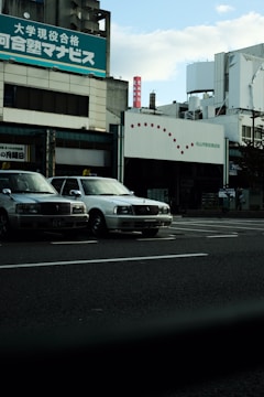 Several taxis are parked alongside a street in front of a commercial building with signage in Japanese. The sky is partially cloudy, and the overall setting gives an urban feel.